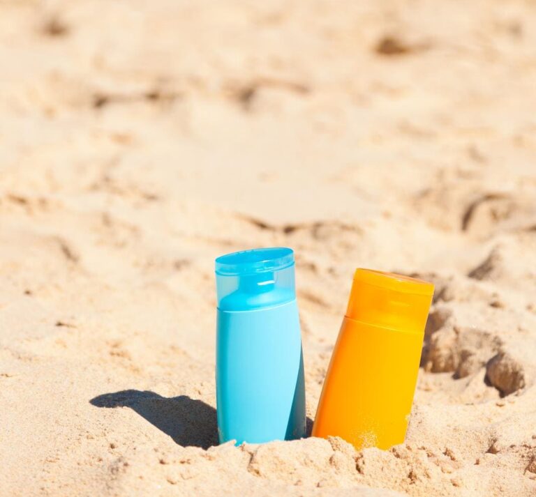 Two sunscreen bottles, one blue and one orange, placed in the sand on an Australian beach with the ocean in the background, illustrating how to avoid sunburn on Australian beaches.