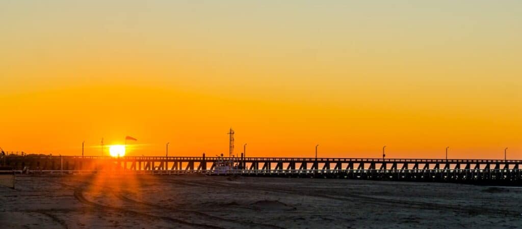 Breathtaking view of sunset in Busselton Jetty with a glowing orange sky silhouetting the long, wooden jetty, which stretches into the sea. This iconic jetty, captured at sunset, adds a dramatic backdrop to the serene beach setting, emphasizing its significance and beauty as a popular tourist destination.