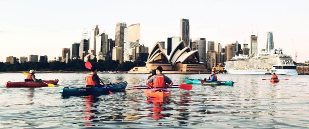 Group on a Sydney kayaking tour, paddling in front of the Sydney Opera House with the city skyline in the background during golden hour.