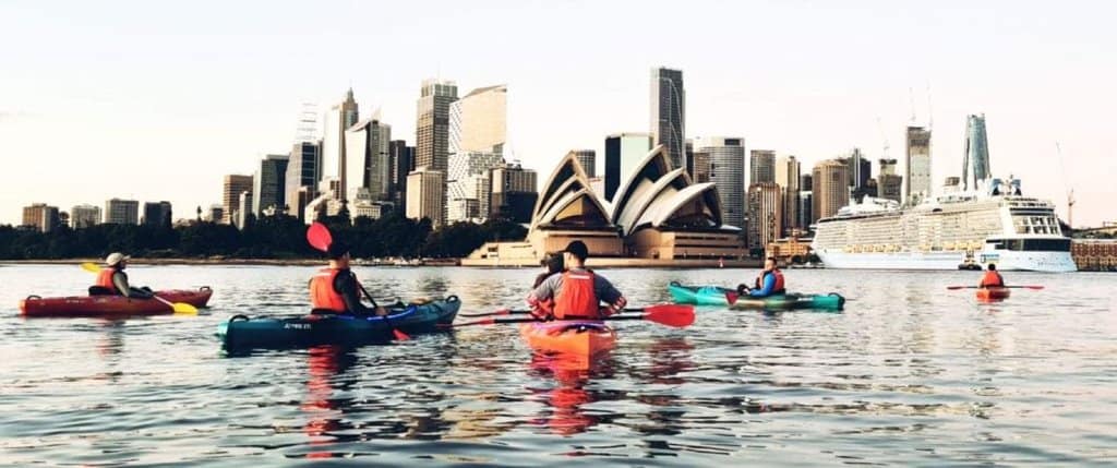 Group on a Sydney kayaking tour, paddling in front of the Sydney Opera House with the city skyline in the background during golden hour.