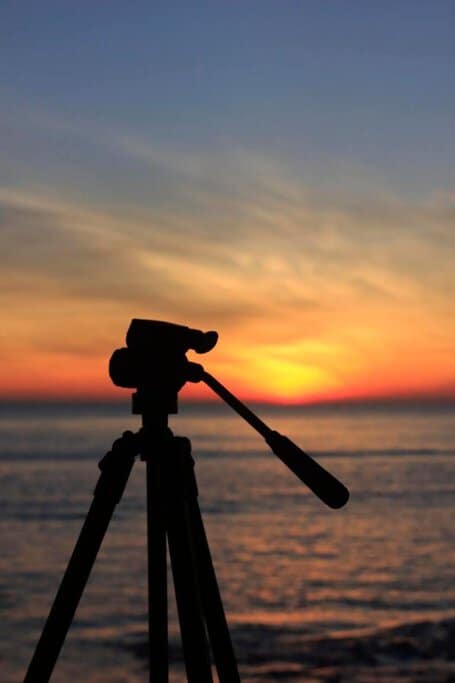 Silhouette of a tripod-mounted camera capturing the vibrant sunset over the ocean, inviting photographers to take some pictures of the breathtaking view.
