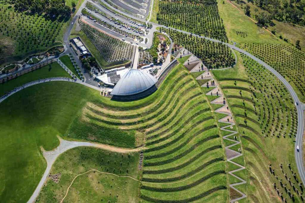 Aerial view of the National Arboretum in Canberra, featuring lush green terraced landscapes, geometric garden designs, and a modern visitor centre. This scenic spot is one of the hidden gems in the Australian Capital Territory (ACT), offering breathtaking views, peaceful walking trails, and a connection to nature. A must-visit for travellers seeking unique outdoor experiences in Canberra.