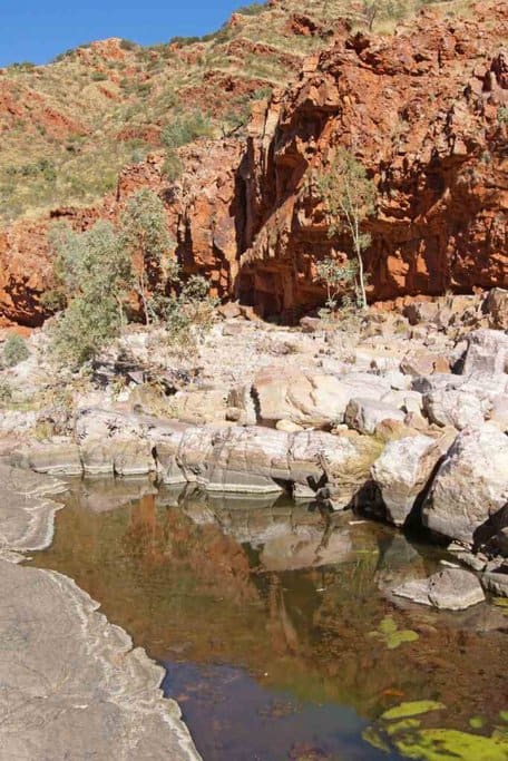 Rocky landscape at West MacDonnell National Park, featuring red cliffs and a reflective waterhole under a clear sky. This image captures the rugged beauty of Australia's outback in summer, highlighting a must-see destination for nature lovers and adventurers.