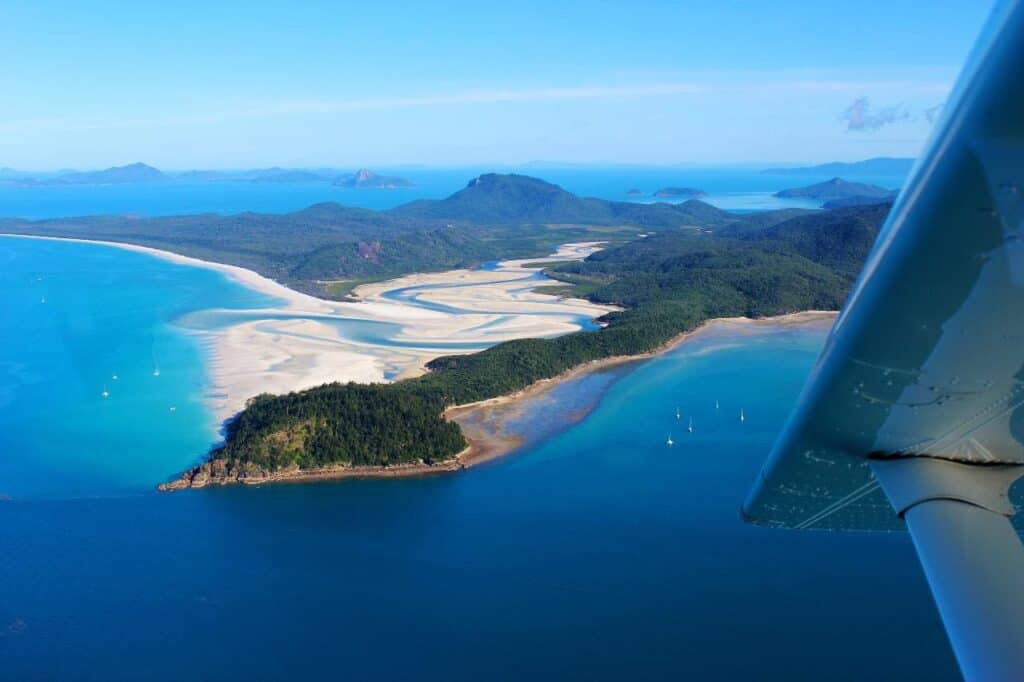 Aerial view from a Whitsunday scenic flight showcasing the stunning Hill Inlet with its dynamic sand patterns blending into the turquoise waters, surrounded by lush greenery and the deep blue sea.